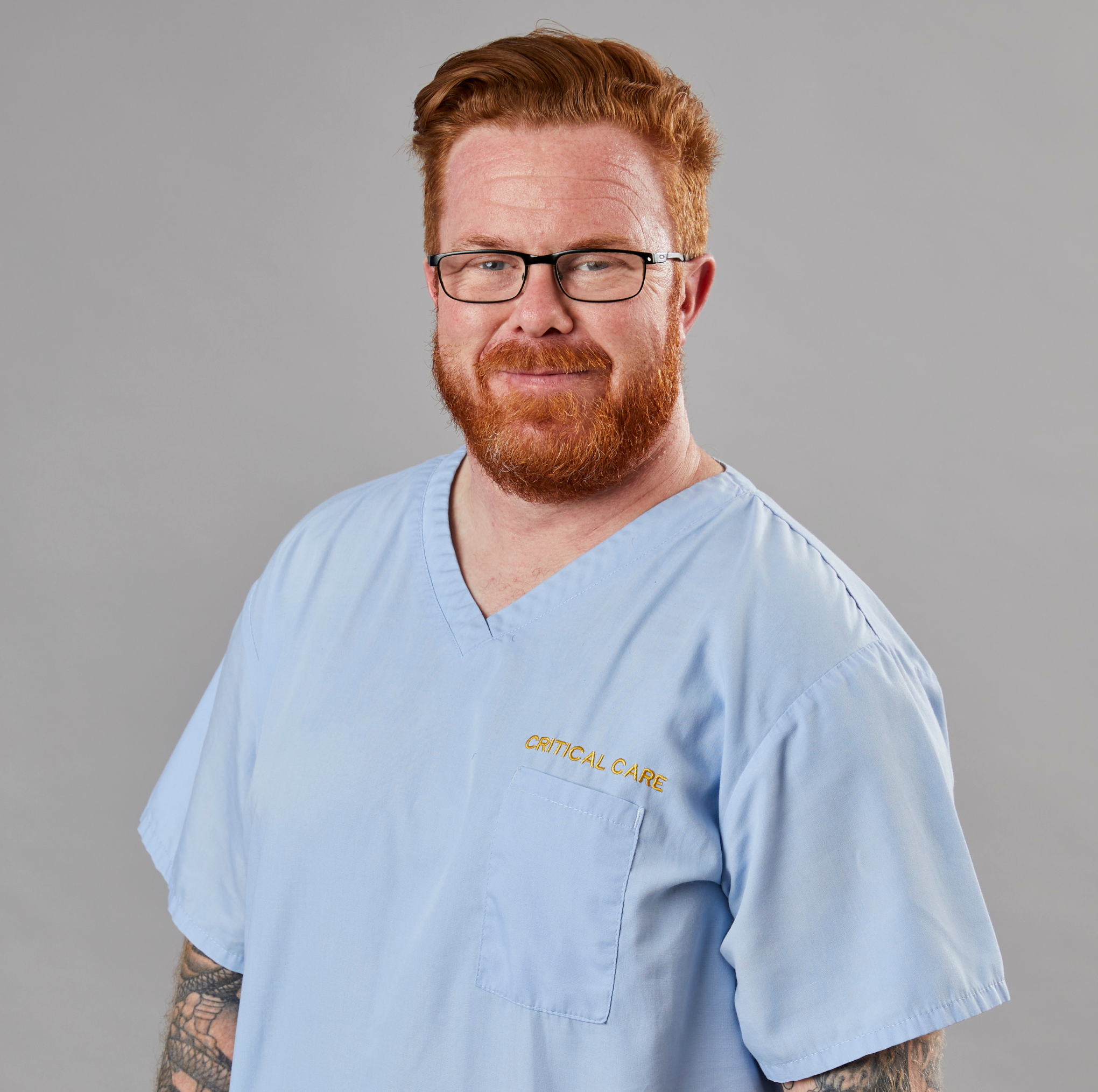 Bearded man in "critical care" scrubs with tattoo sleeves, smiling.