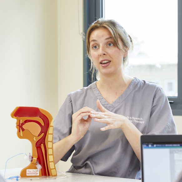 Speech therapist in gray scrubs explaining with hand gestures beside a vocal tract model.