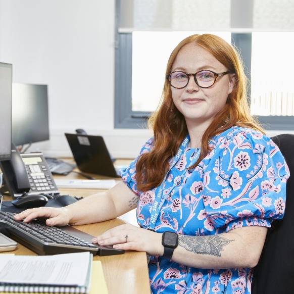 Woman with red hair and glasses working at a desk, tattoo visible on left arm.
