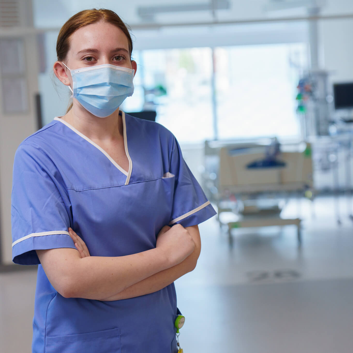 Female healthcare worker in blue scrubs and mask standing with arms crossed in hospital.