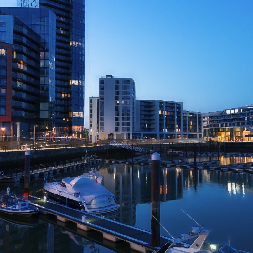 Twilight marina with boats, modern buildings, and reflections on calm water.