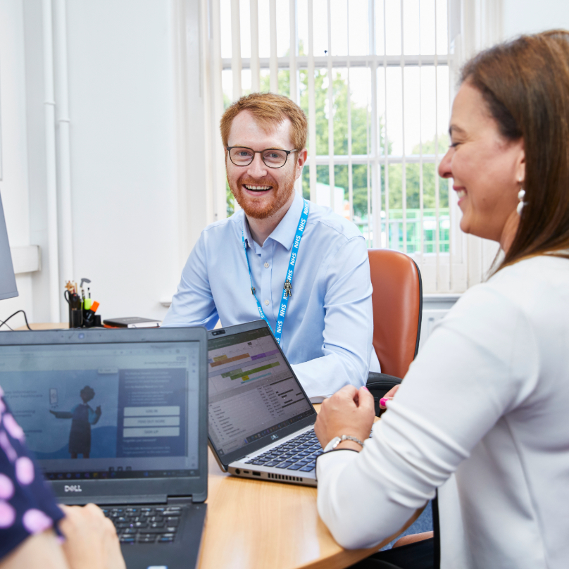 Two colleagues with laptops talking in bright office with whiteboard.