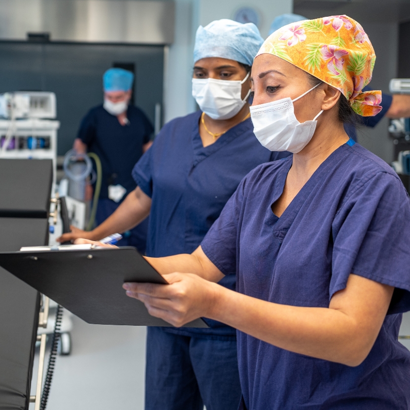 Two masked healthcare workers in scrubs reviewing a clipboard in hospital.