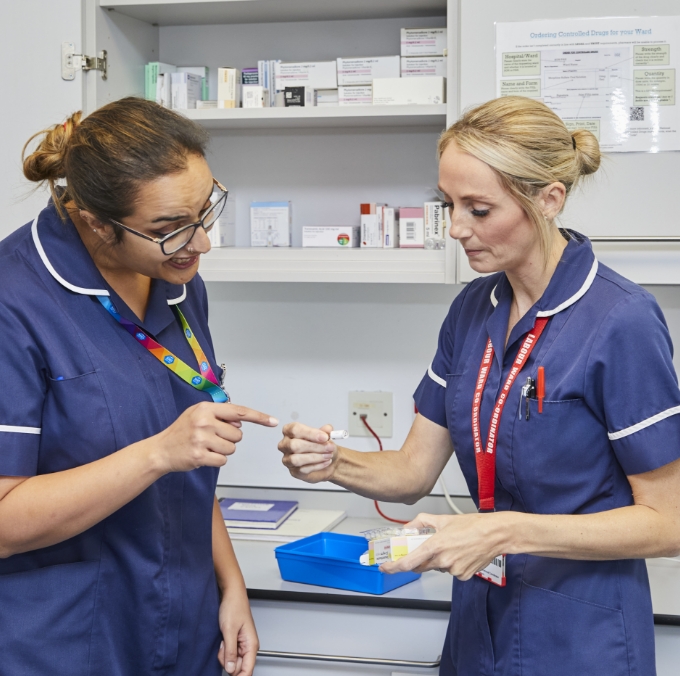 Two nurses discussing a syringe in a hospital supply room.