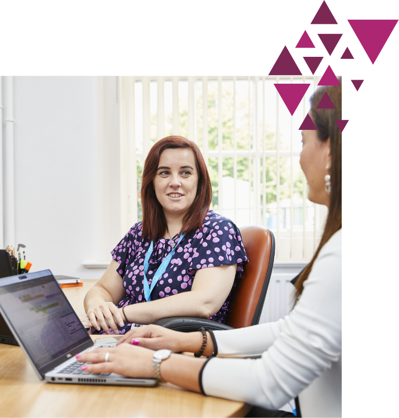 Two women smiling and talking at a desk with a laptop in a bright office
