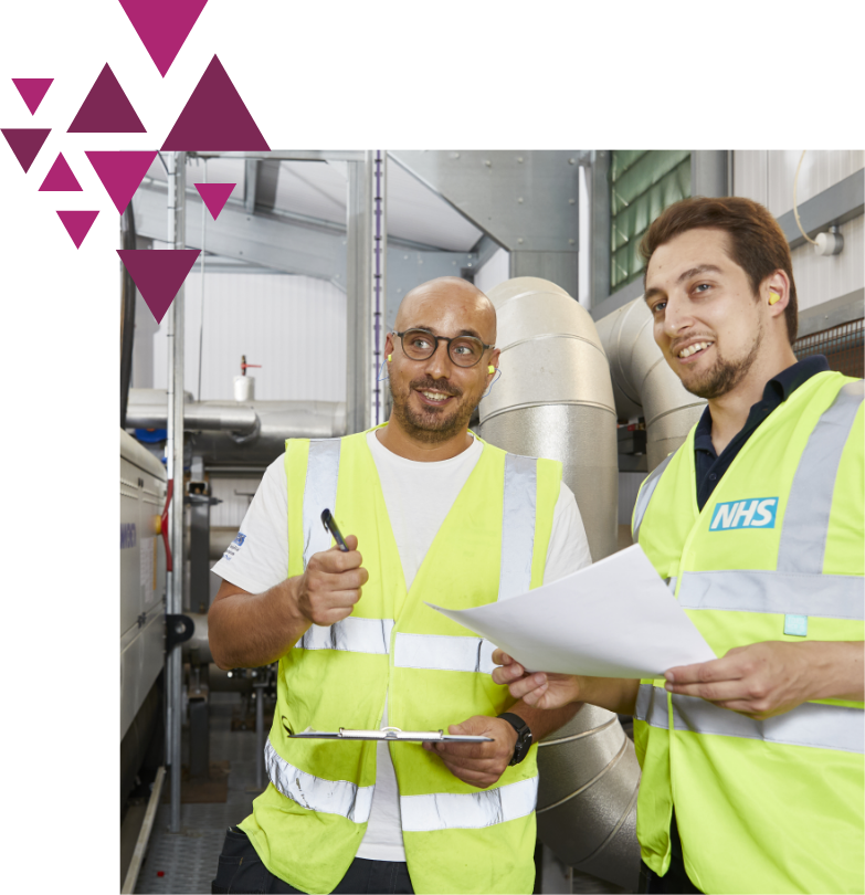 Two men in NHS safety vests discuss paperwork in industrial setting