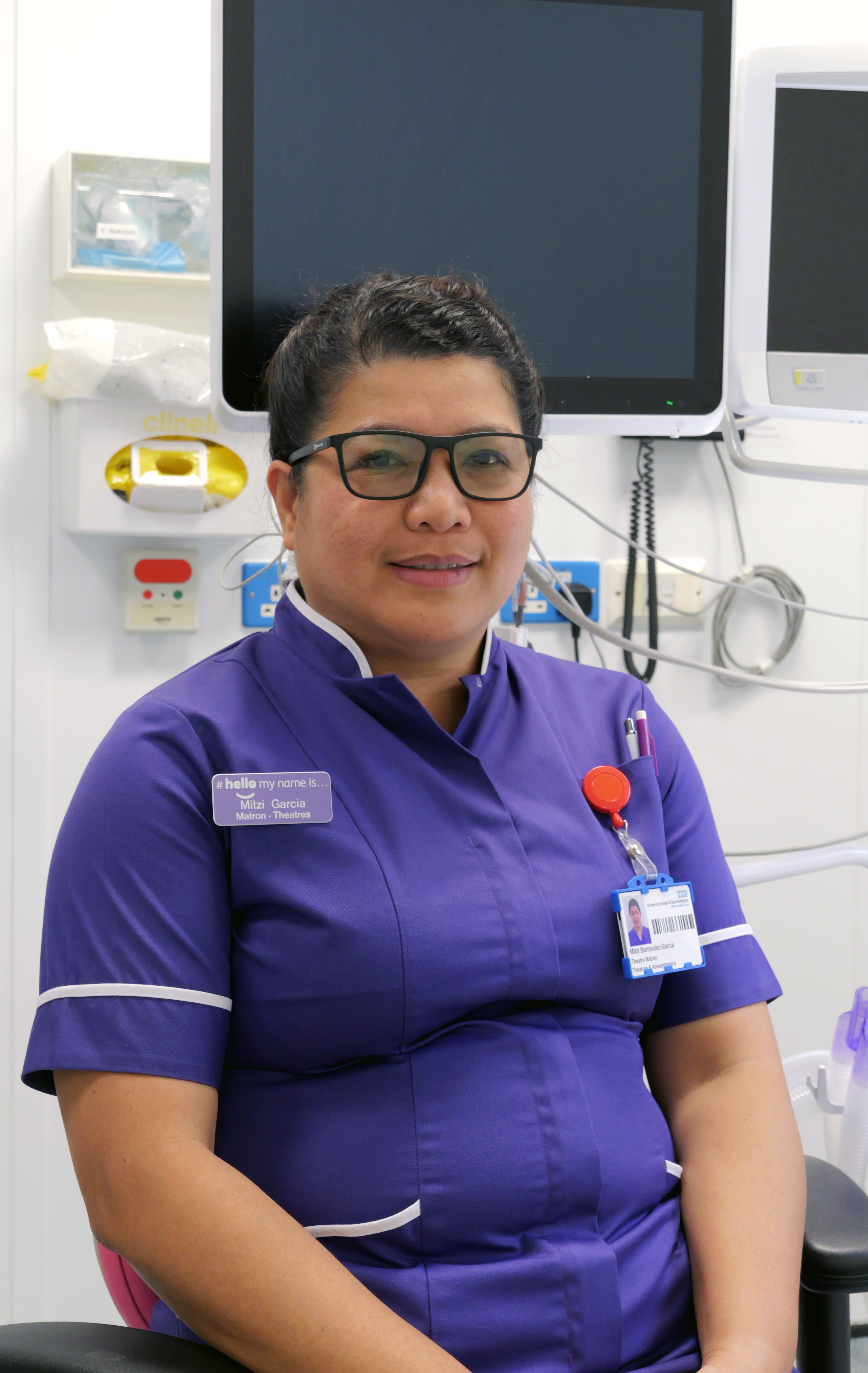Smiling healthcare worker in purple uniform with glasses, seated in a clinical room.