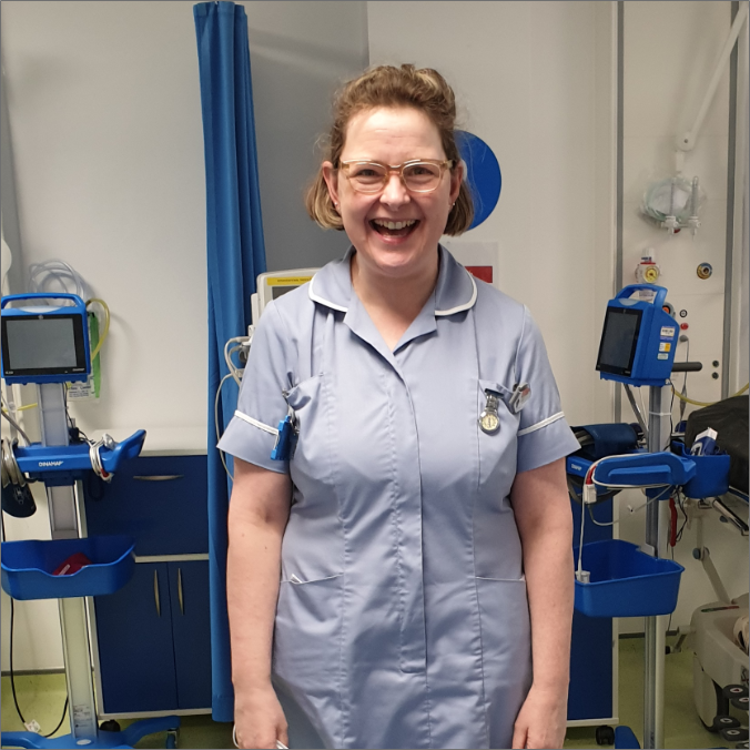 Smiling nurse in pale blue uniform in hospital room with medical equipment.