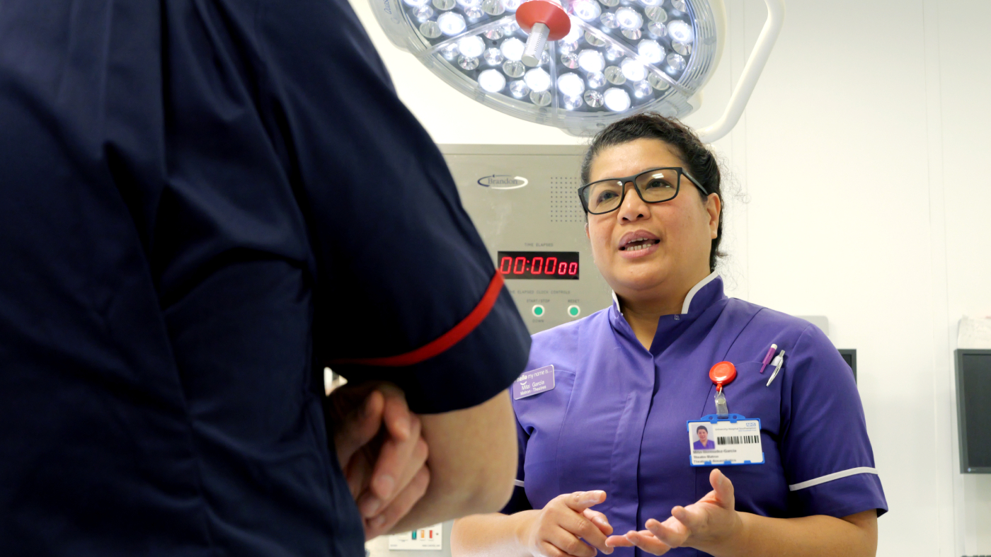 Nurse in purple scrubs talks with a colleague under surgical lights.