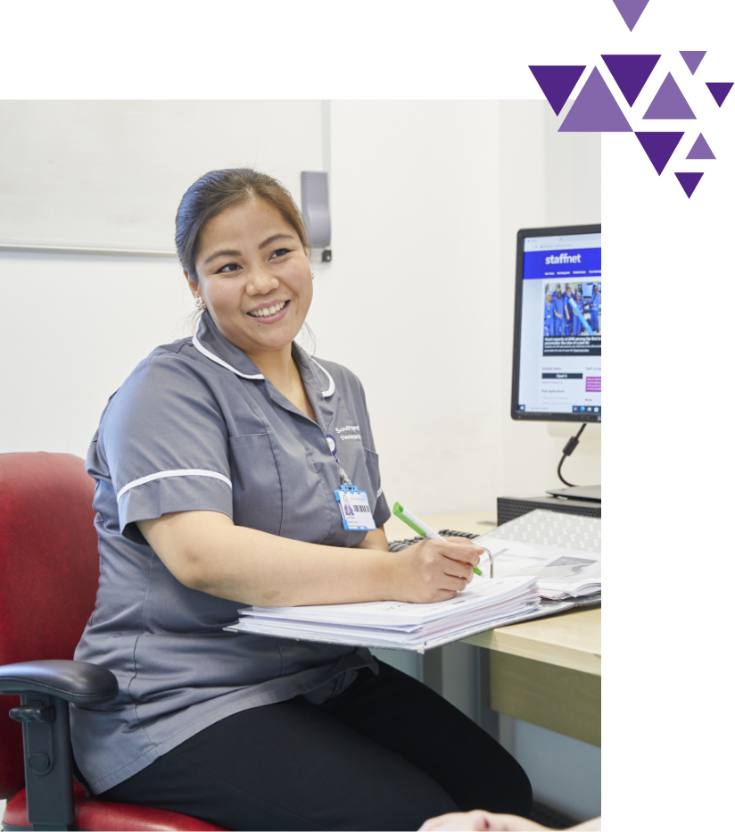 Smiling nurse in scrubs writing at desk with computer and purple graphics