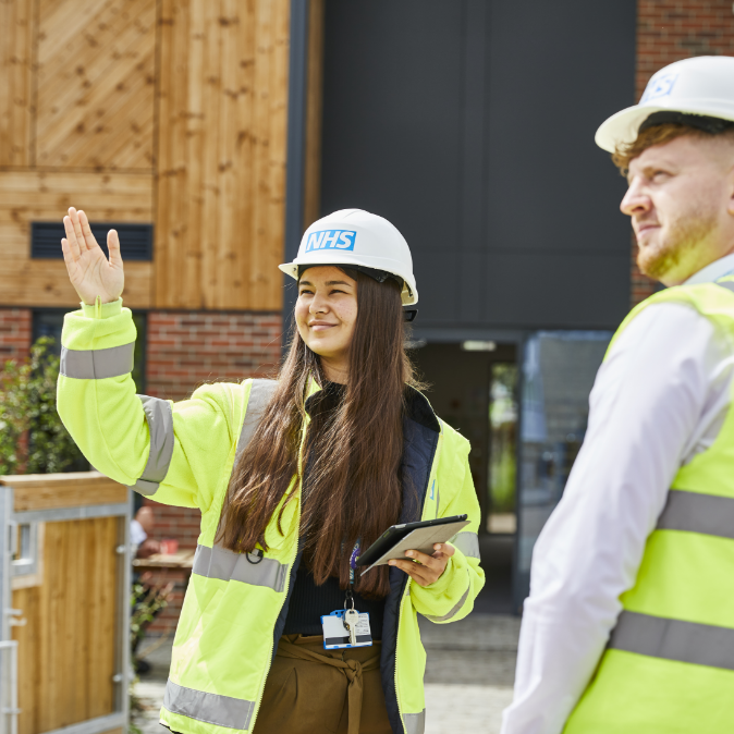 Female construction worker in NHS gear gestures with tablet outside building