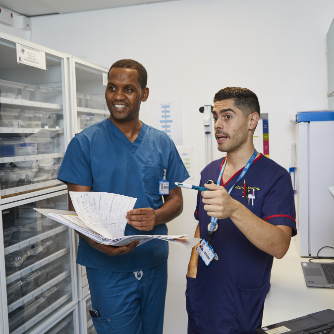 Two male healthcare workers in scrubs discussing documents in supply room